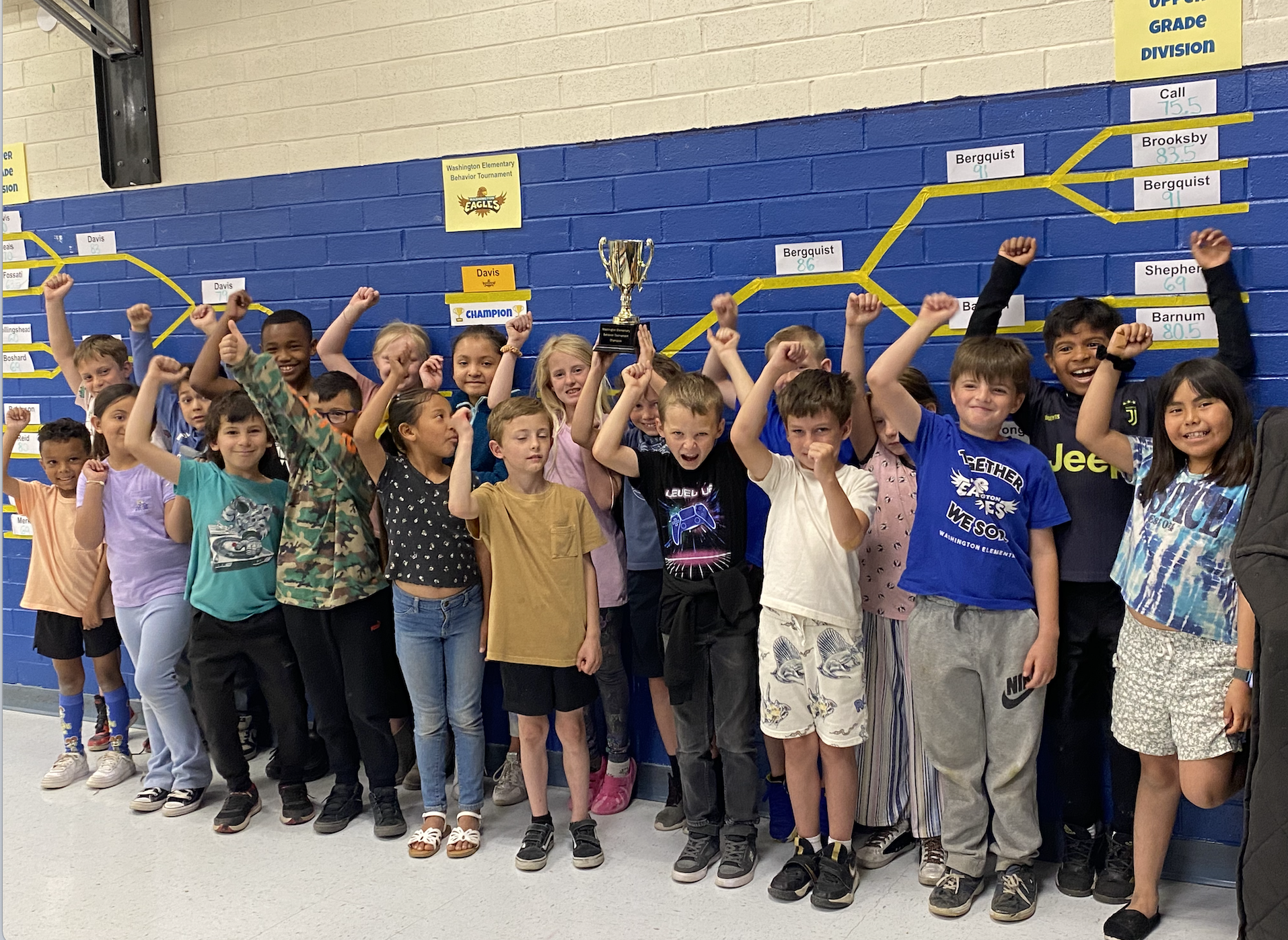 Mrs. Davis's class is standing in front of the behavior tournament bracket in the lunchroom holding up the championship trophy